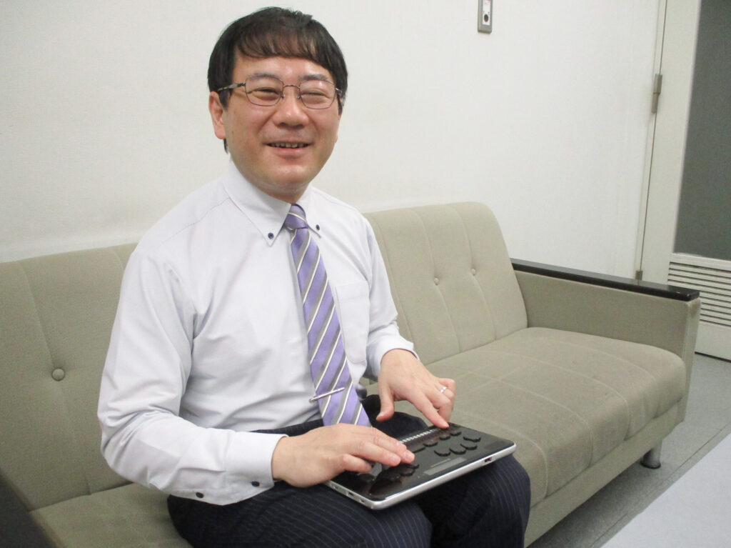 写真：佐木理人さん。膝に点字ディスプレイをのせた笑顔の男性